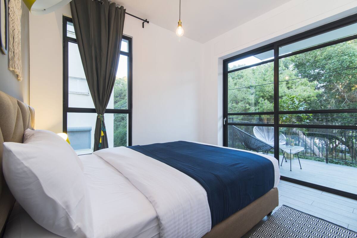 A bedroom with a large bed, a view of the outdoors through a window, and a balcony with chairs.