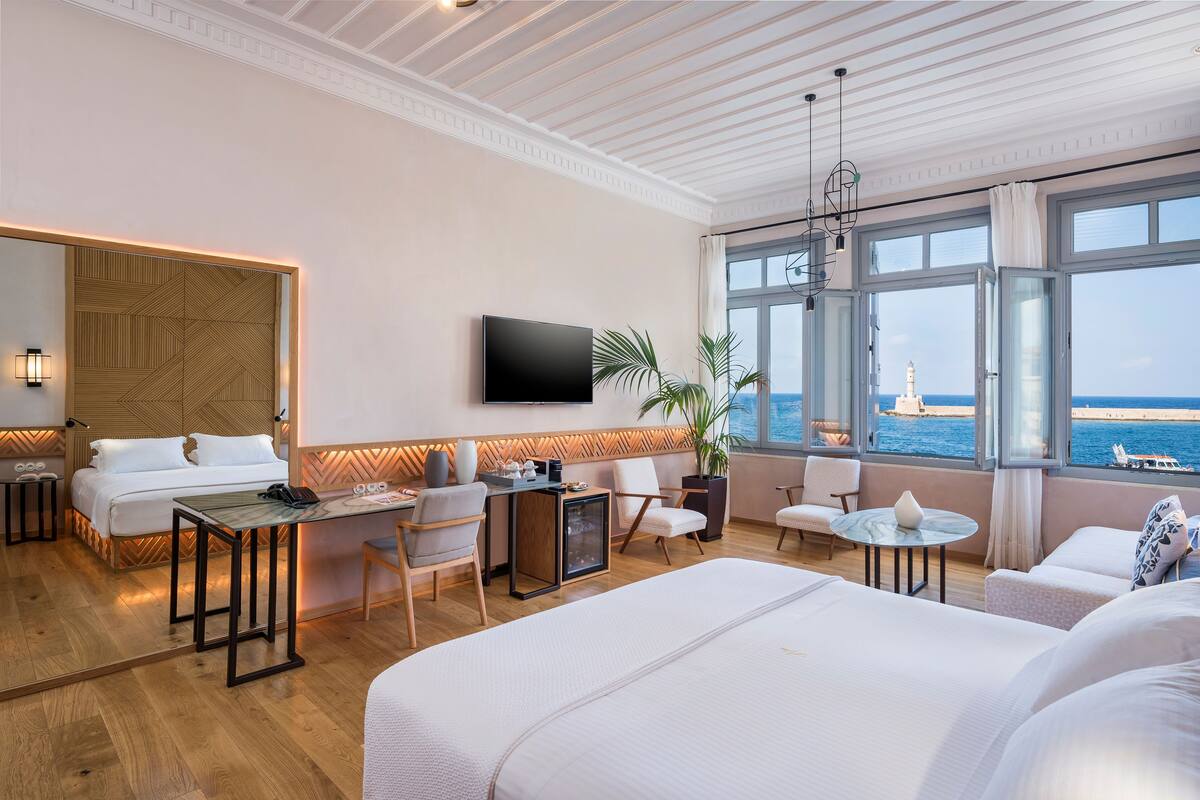 A hotel room with a large bed, a desk, a chair, a small table, and a TV mounted on the wall. There is a large window with a view of the sea and a lighthouse.