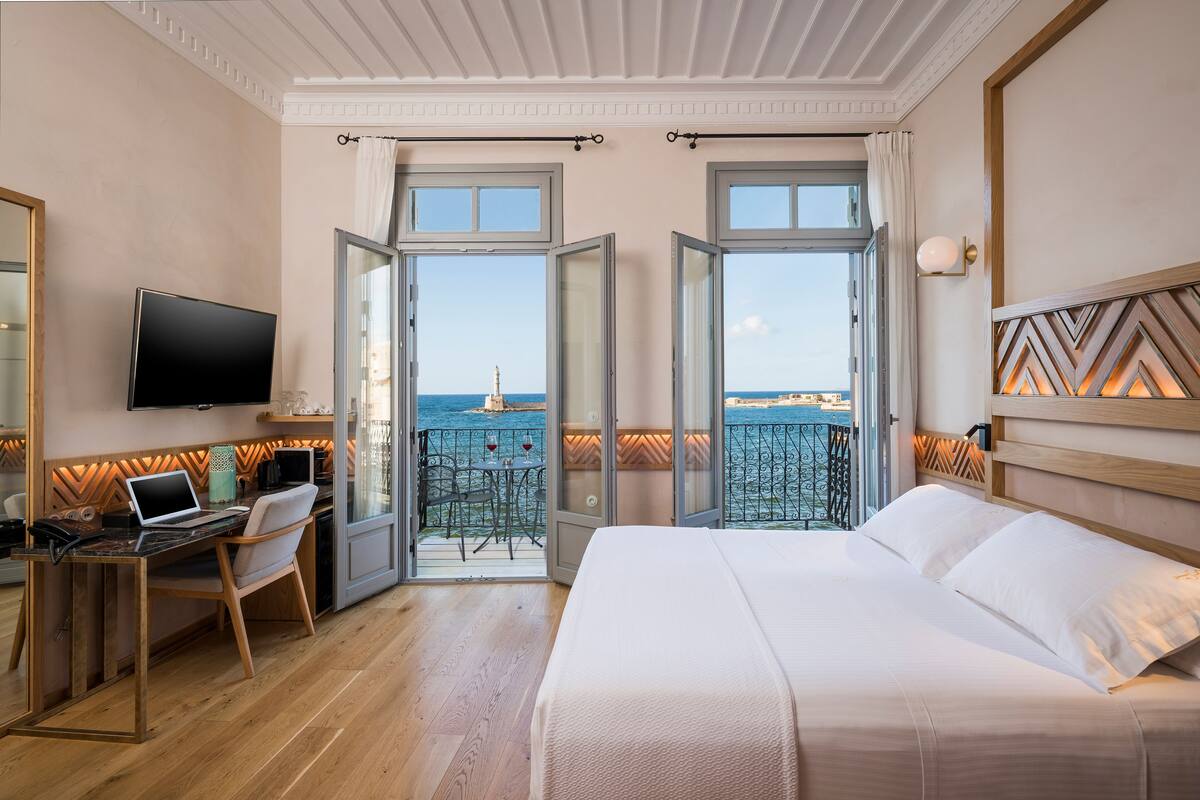 A hotel room with a bed, a desk with a laptop, a TV, and a balcony with a view of the sea.