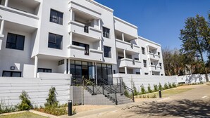 Un edificio residencial moderno de varios pisos, con balcones, una entrada central y un jardín delantero ajardinado.
