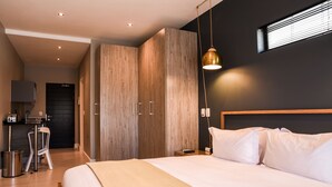 Una habitación de hotel moderna con una cama grande, un armario de madera, una pequeña kitchenette y una ventana con persianas.