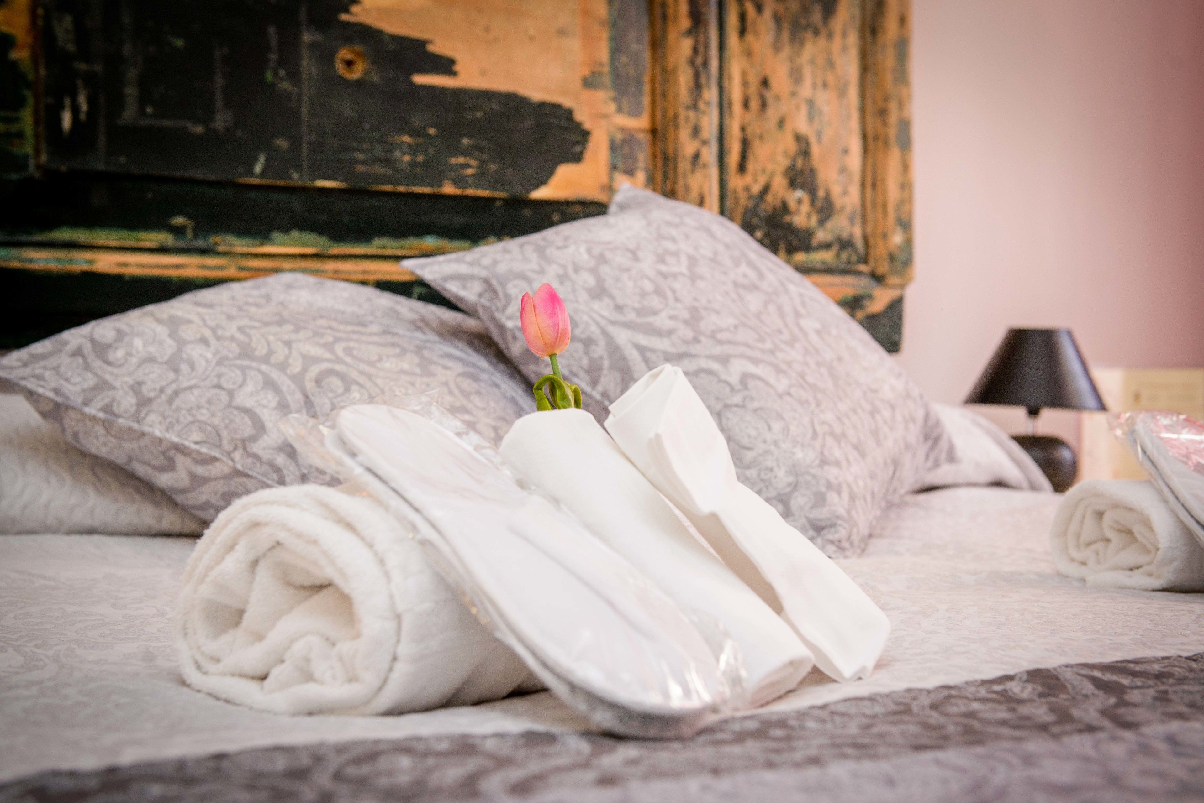 Una cama bien hecha con toallas blancas enrolladas y una flor rosa, sobre un fondo de madera rústica.