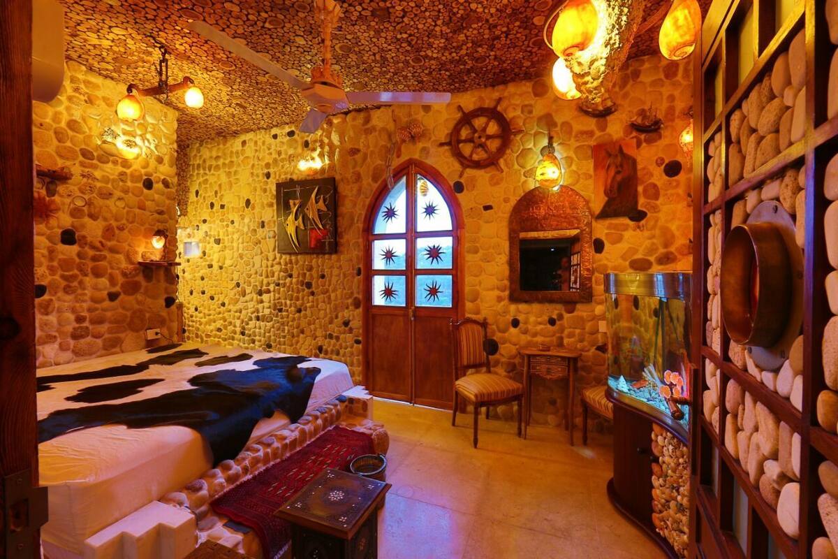 A stone-walled room with a bed, a wooden chair, a fish tank, and a decorative wall with a ship's wheel.