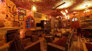 Una habitación acogedora y rústica con paredes de piedra, muebles de madera y chimenea. Hay varias zonas de estar, incluyendo un sofá y sillones, con una alfombra estampada en el suelo.