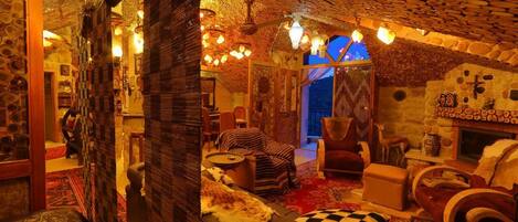 A cozy room with a stone ceiling, wooden walls, and a fireplace. There are several seating areas with cushions and a large window providing natural light.