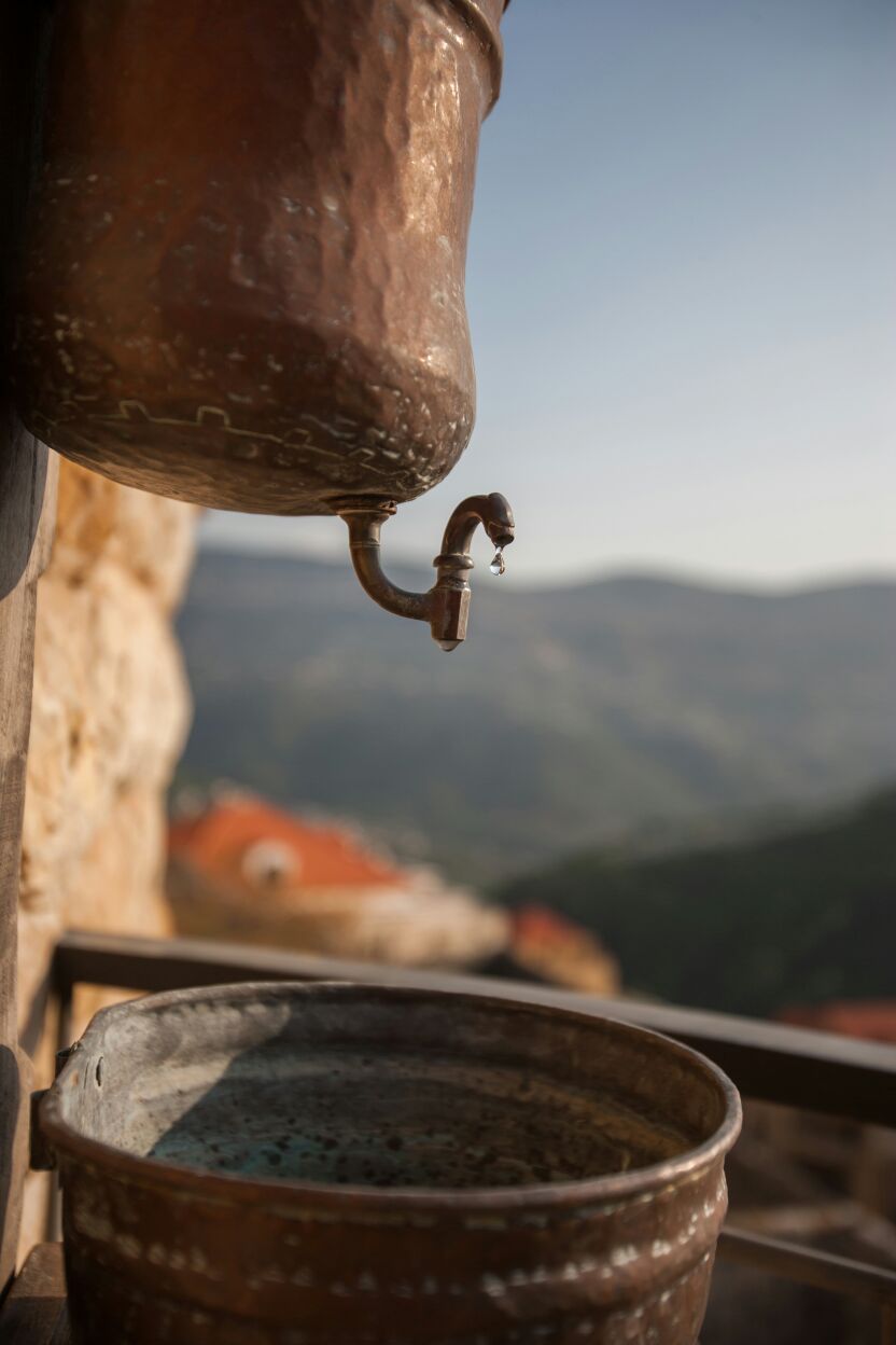 A rustic metal container with a spout, likely used for water, placed on a ledge with a mountainous backdrop.