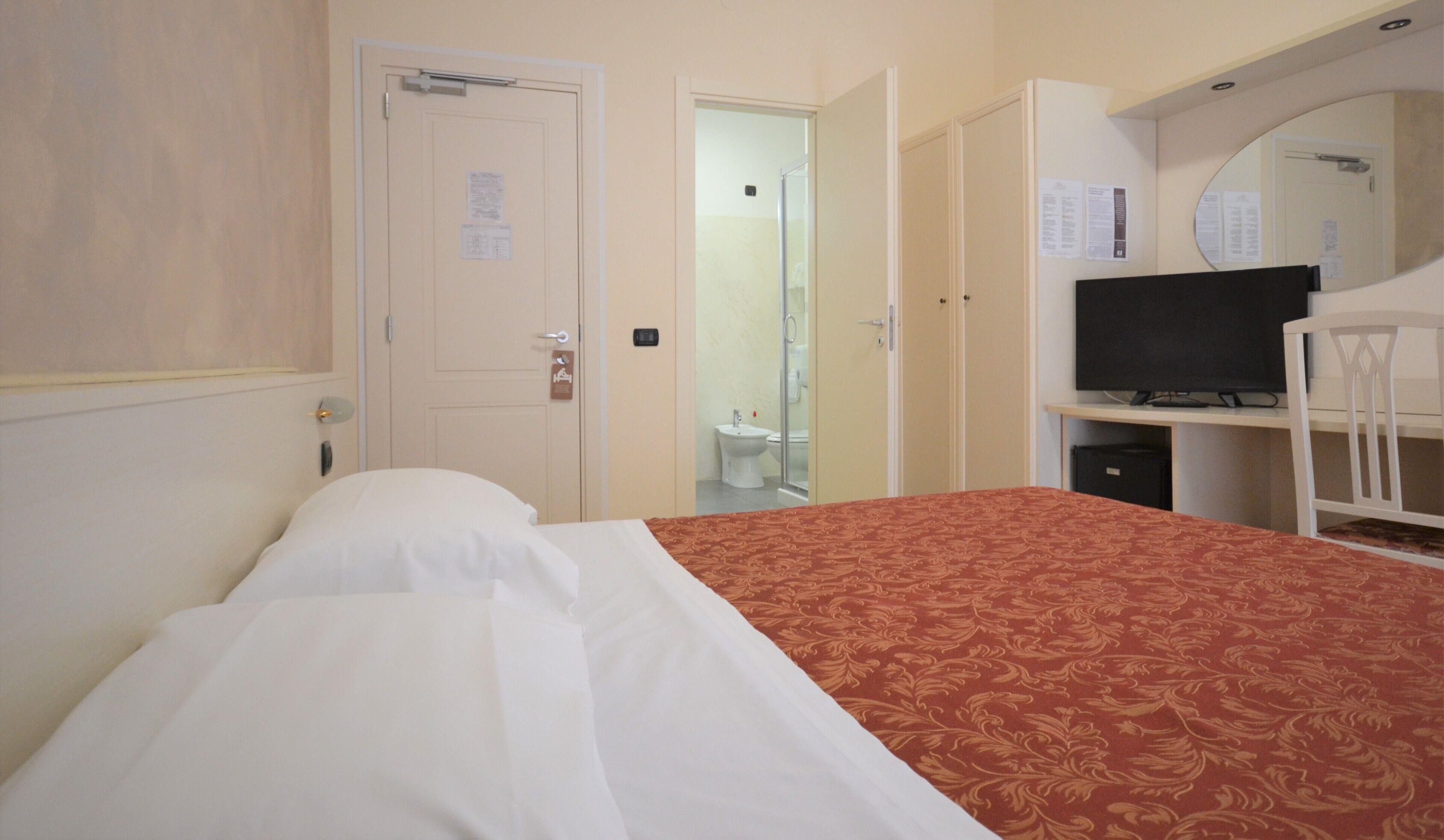 Quarto de hotel com uma cama, uma escrivaninha com televisão, um espelho e um banheiro visível através de uma porta aberta.