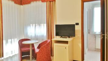 A hotel room with a television on a stand, a small round table, two red chairs, and a door leading to a bathroom.