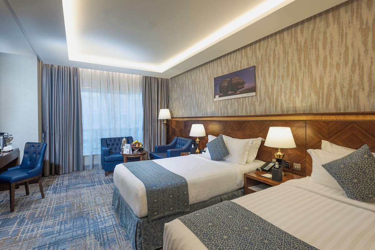 A hotel room with two beds, a desk, chairs, and a large window with curtains.