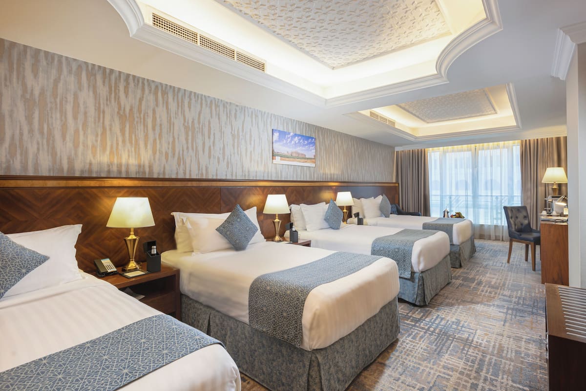 A hotel room with three beds, a desk with a lamp, and a view of the city.