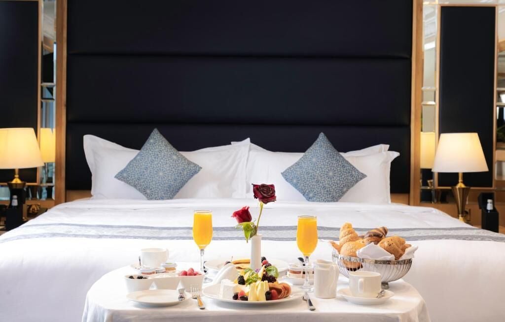 A neatly made bed with a dark headboard, white pillows, and a breakfast tray with fruits, pastries, and drinks.