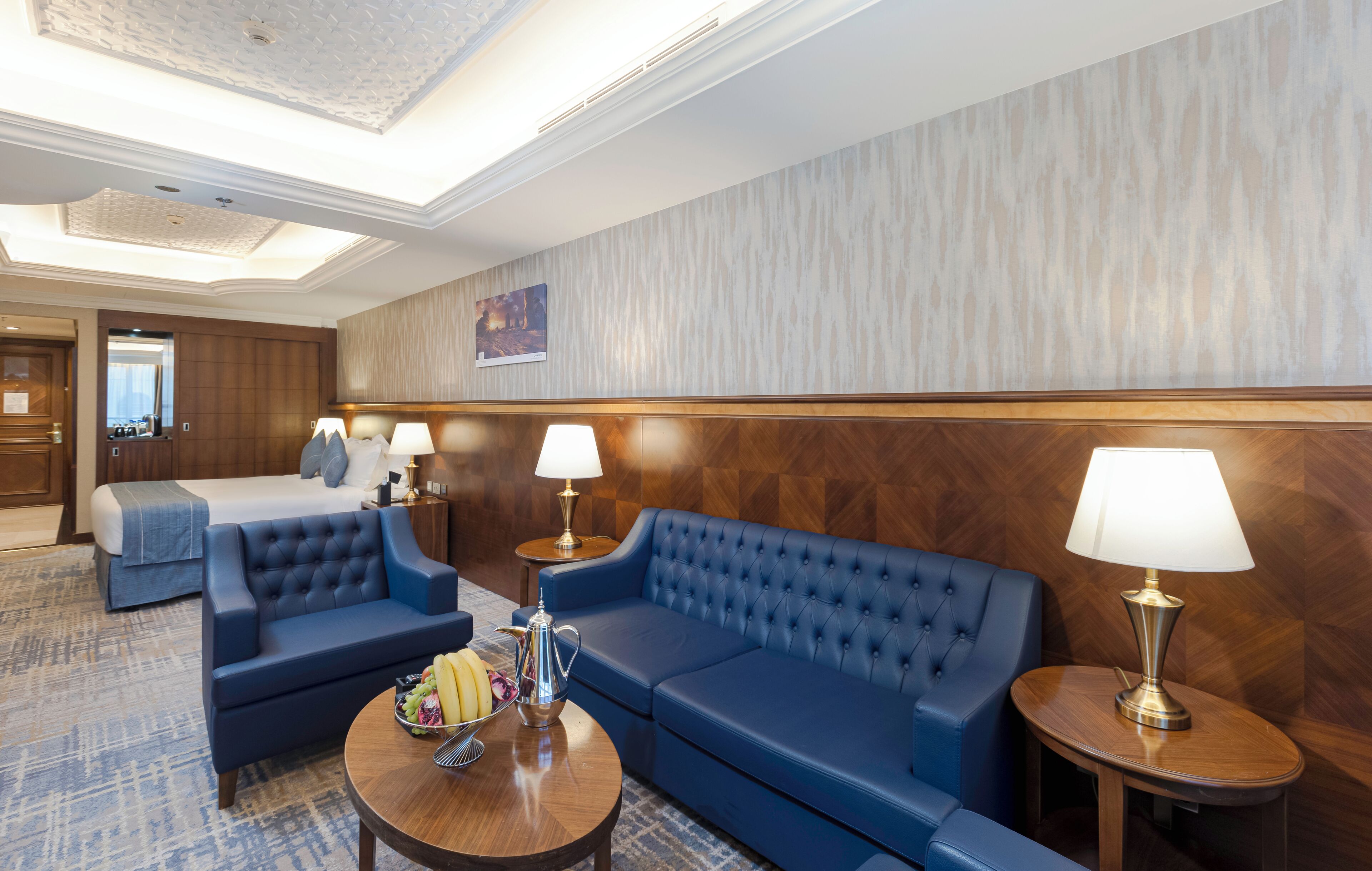 A hotel lobby with a blue sofa set, a round coffee table with fruits and a teapot, and two lamps on wooden side tables.