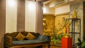 A wooden sofa with cushions, a golden statue, and a red pedestal base in a room with wooden flooring and a stone wall. - Porwa House (Chiang Mai)