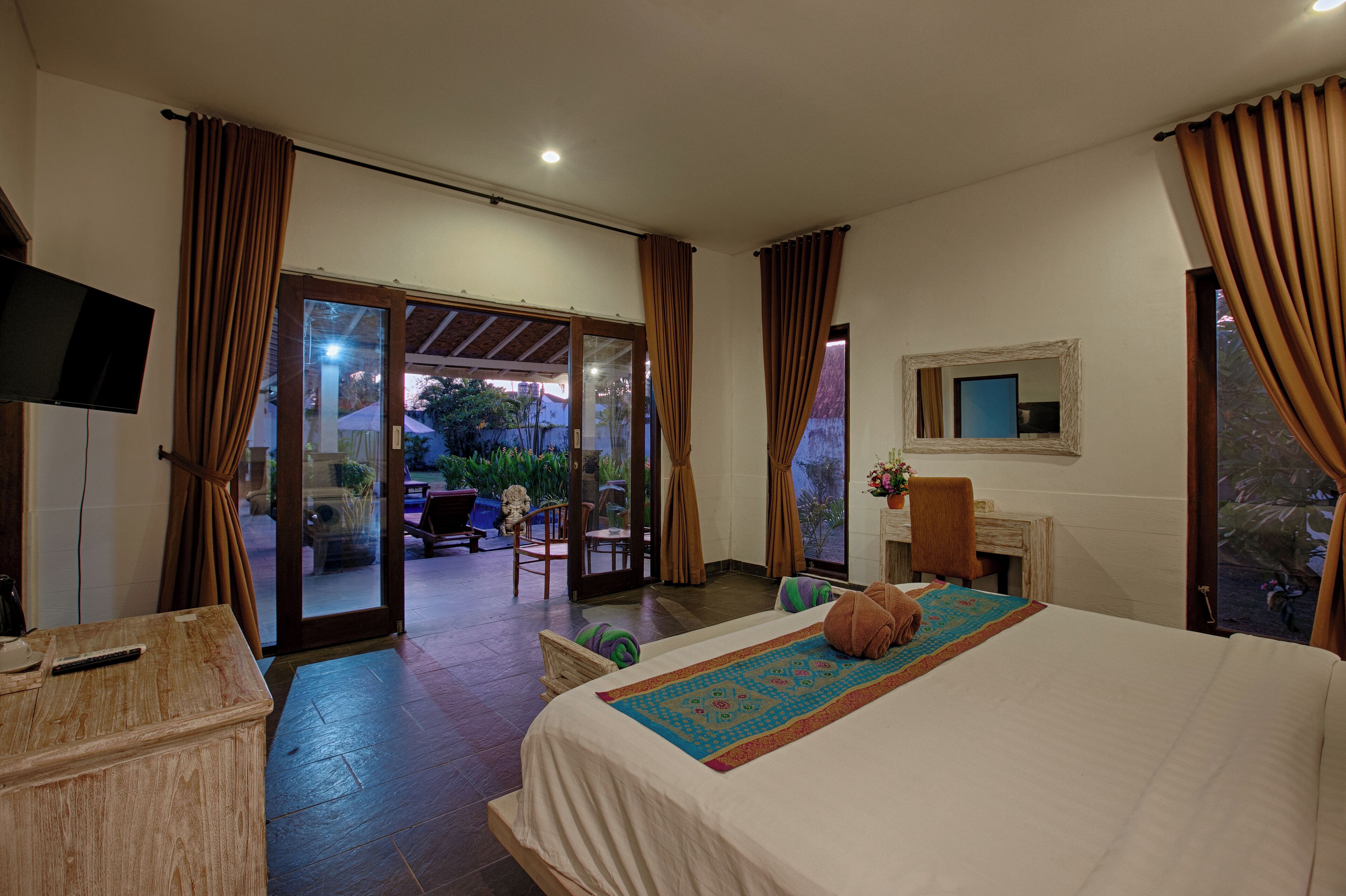 Habitación de hotel con cama, escritorio, silla, televisor y puerta de vidrio corredera que da a un espacio al aire libre.