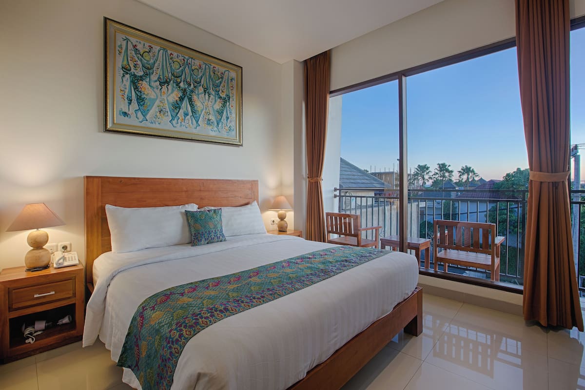 Une chambre d’hôtel avec un grand lit, une tête de lit en bois, deux tables de chevet avec des lampes, une couverture colorée, une grande fenêtre donnant sur la ville et un balcon avec une chaise et une table.