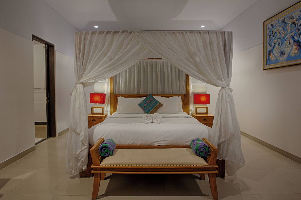 Une chambre d’hôtel bien aménagée, avec un grand lit, deux lampes de chevet, un banc au pied du lit et un tableau au mur.