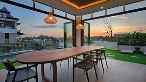 A modern dining area with a long wooden table and chairs, large windows offering views of a residential area and a body of water during sunset.
