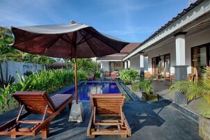 Uma área de piscina com espreguiçadeiras de madeira e um grande guarda-sol, cercada por vegetação exuberante e uma estrutura de telhado em cerâmica.