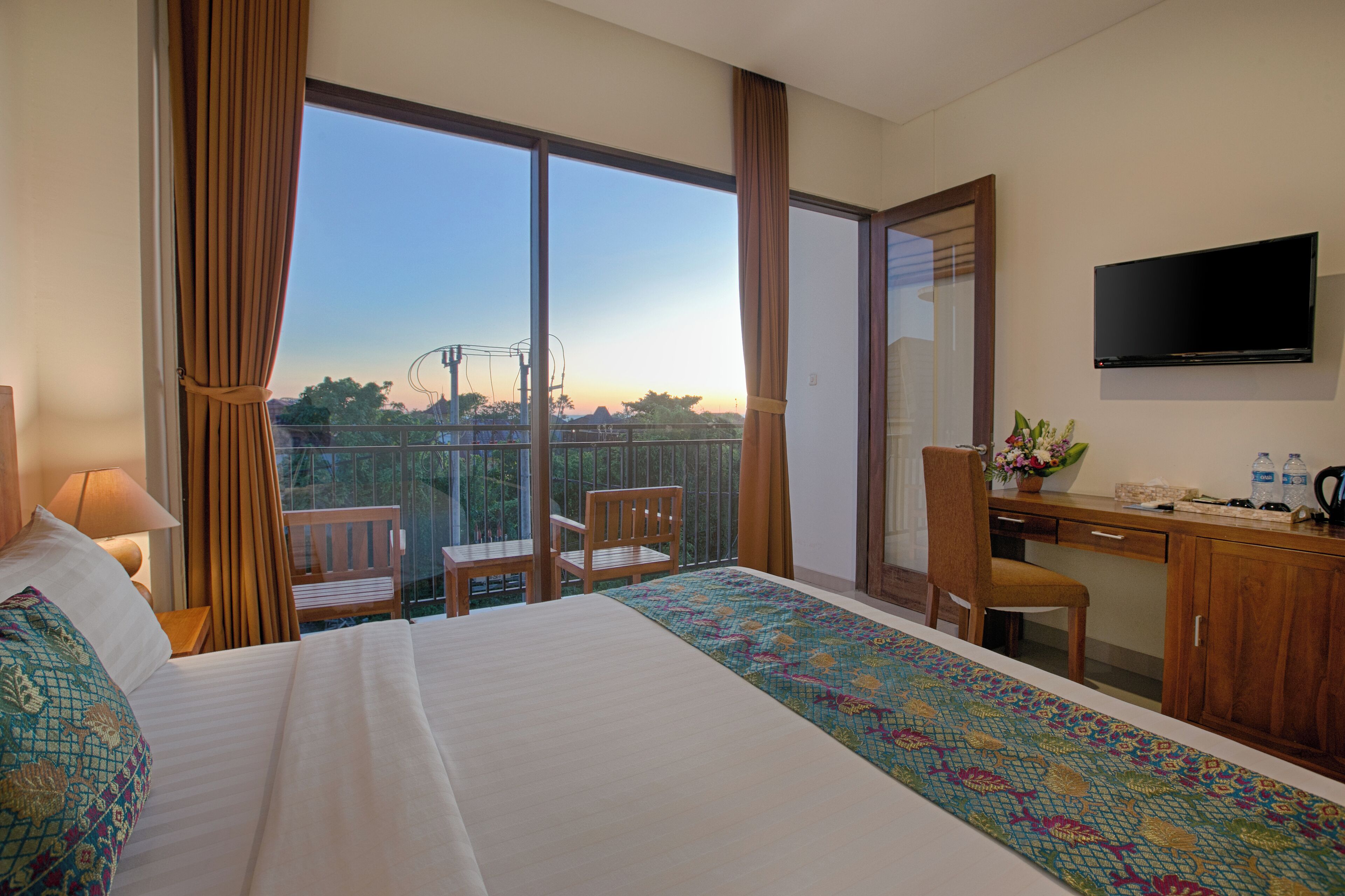 A hotel room with a large bed, a desk with a chair, a TV, and a balcony with a view of the city.