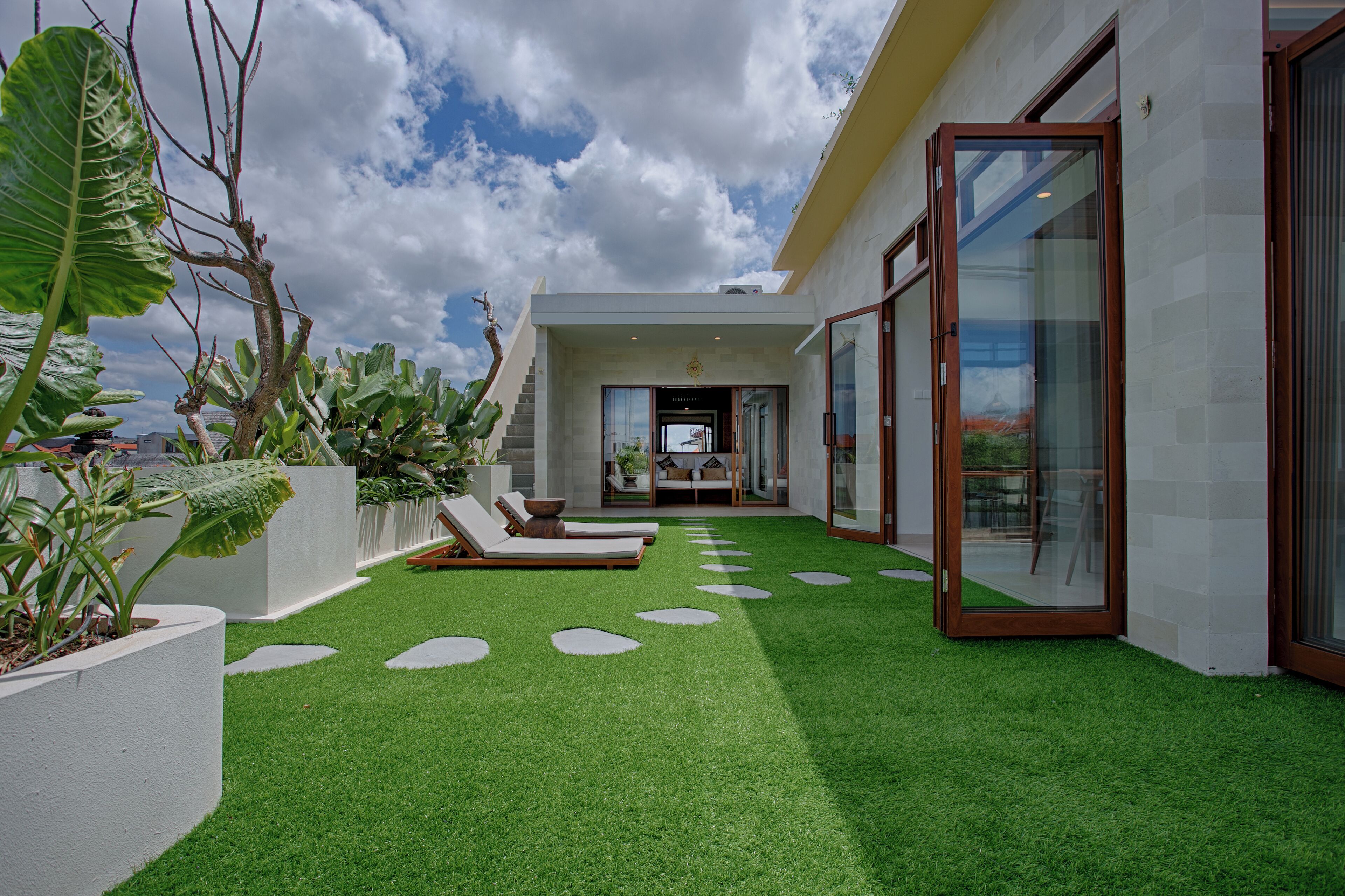 A modern outdoor area with artificial grass, lounge chairs, and a glass door leading to a covered entrance.