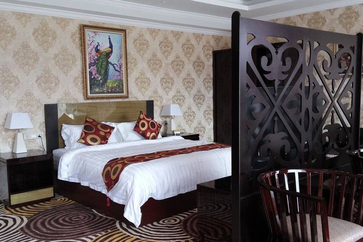 A hotel room with a large bed, two bedside tables with lamps, a decorative headboard, a patterned carpet, and a large window with a view of trees.