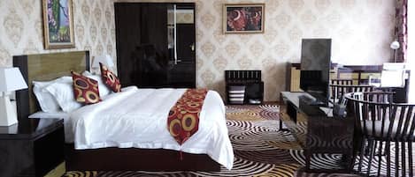 A hotel room with a large bed, a desk, a chair, a TV, and an air conditioning unit.