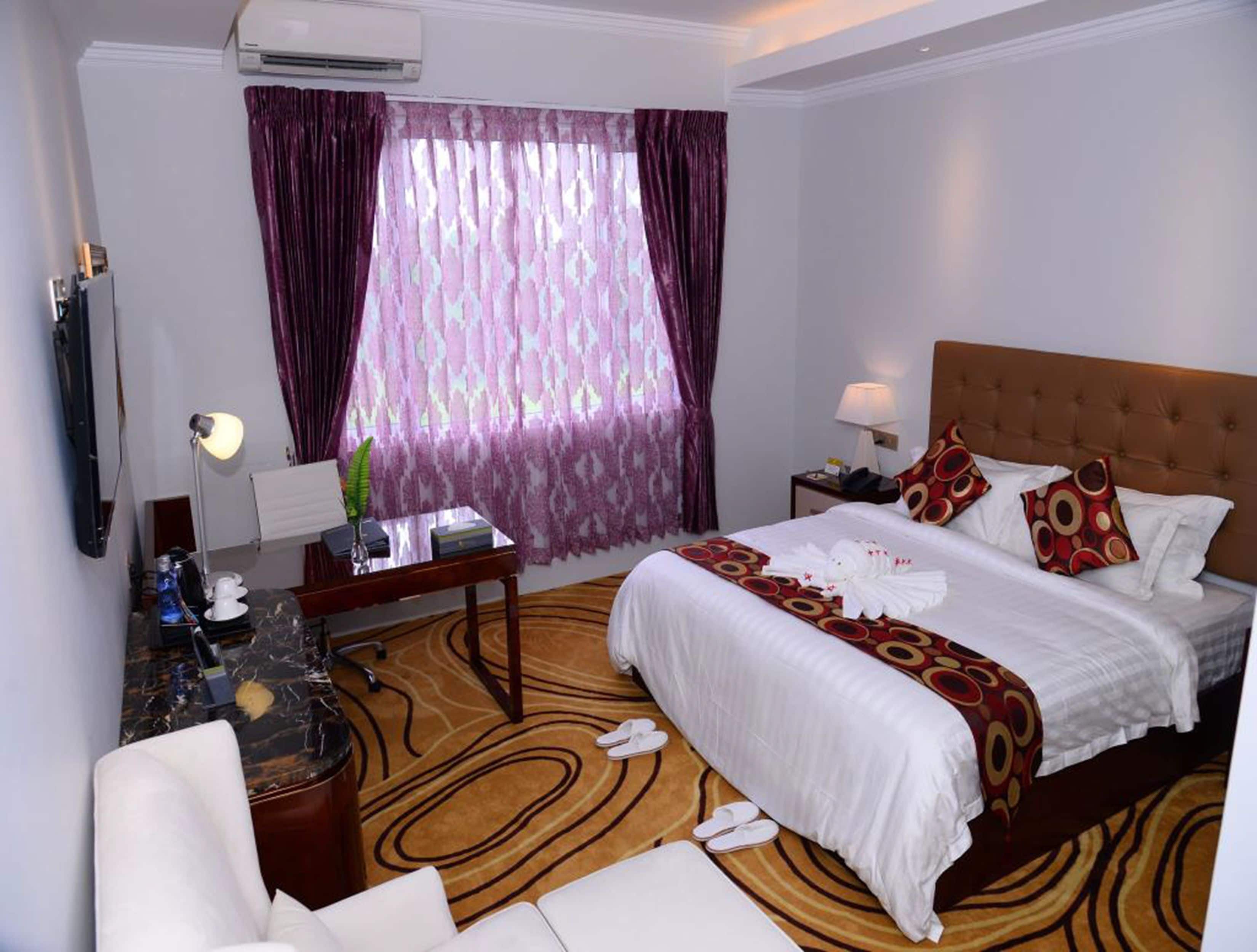 Habitación de hotel con una cama grande, un escritorio con lámpara, una silla, un televisor y una ventana con cortinas.