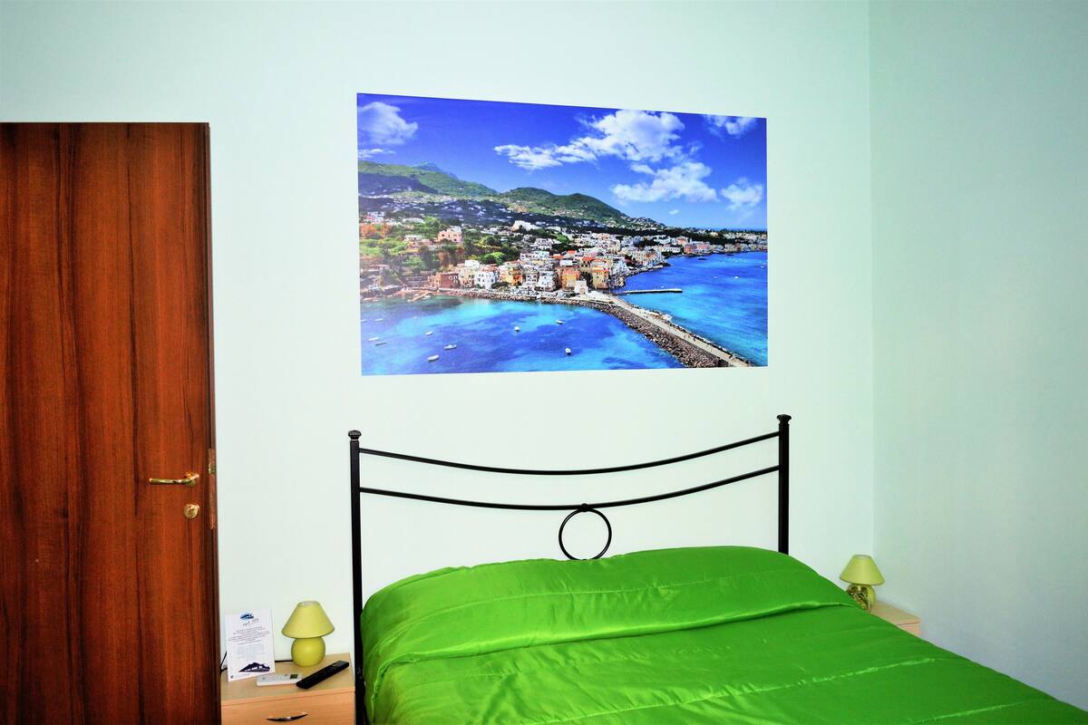 A bedroom with a bed, a wooden door, a nightstand with a lamp, and a wall-mounted picture of a coastal town.