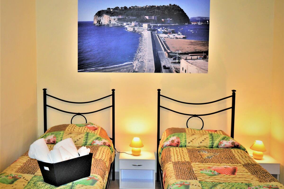 A hotel room with two beds, each with a headboard and a bedside table with a lamp. A framed picture of a coastal scene is on the wall above the beds.