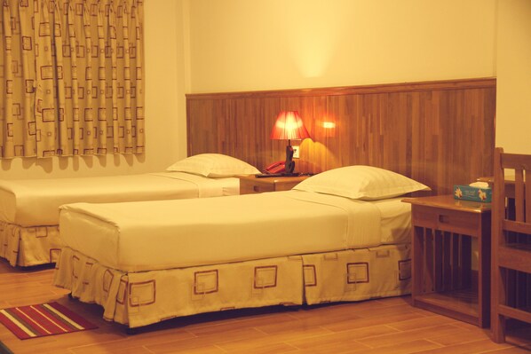 Habitación de hotel con dos camas, un cabecero de madera, una mesita de noche con lámpara y una pequeña mesa con una caja.