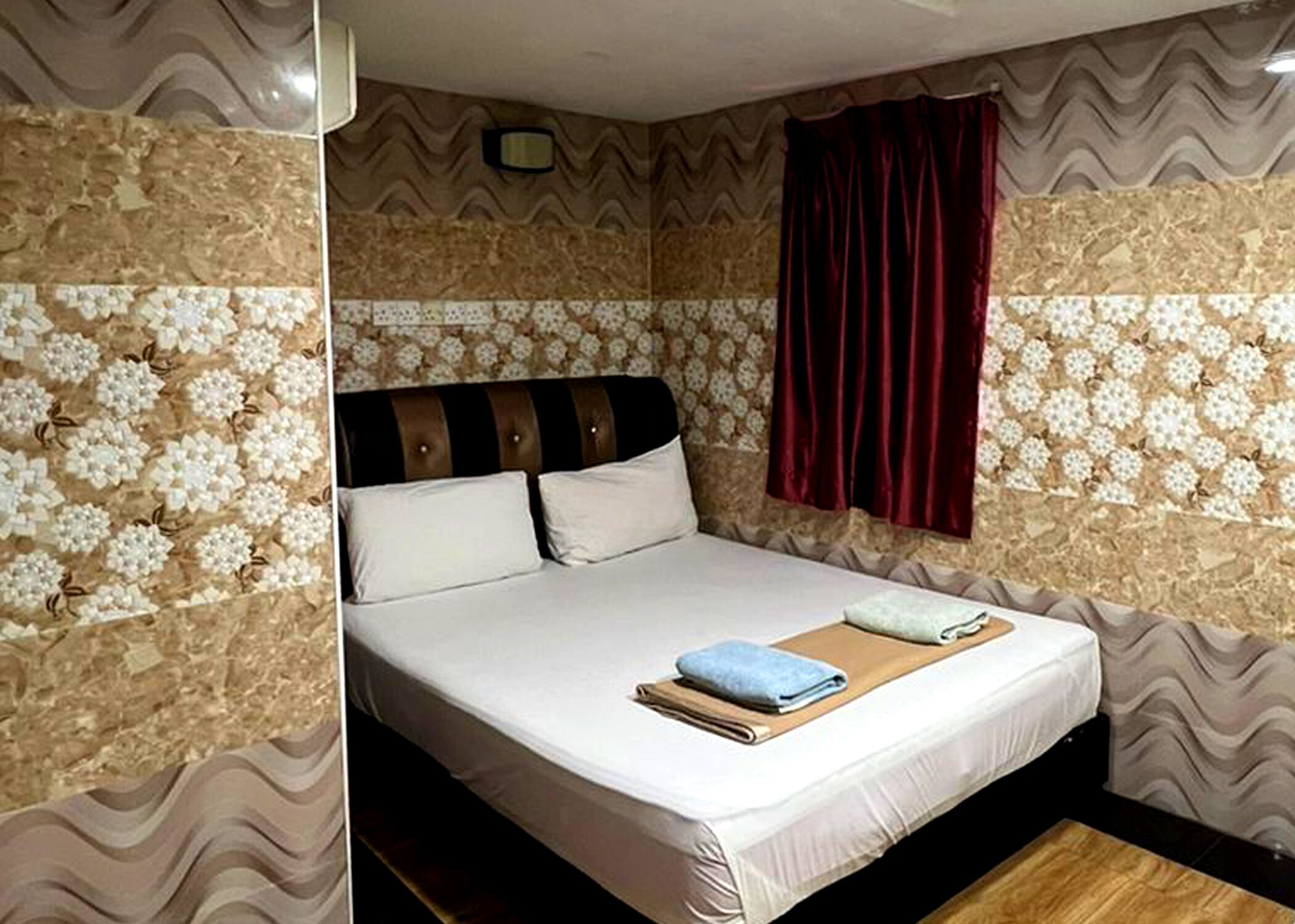 A neatly made bed with a white mattress, two pillows, and a blue towel. The room has patterned wallpaper and a window with red curtains.