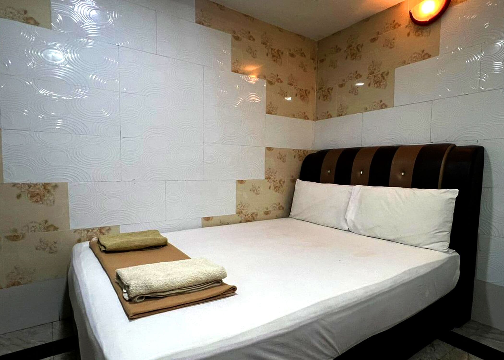 Habitación de hotel con cama, ropa blanca, cabecero a rayas y una lámpara fijada a la pared.