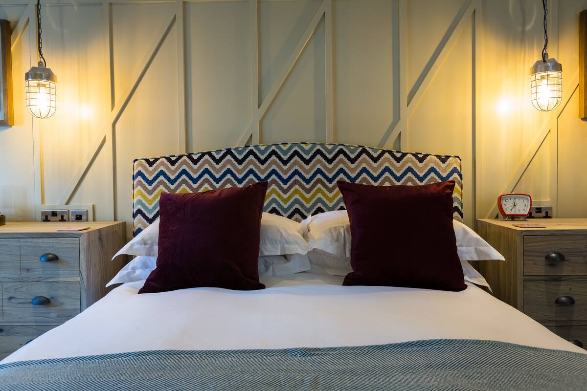 A neatly made bed with a chevron-patterned headboard, two dark pillows, and a bedside table with a red alarm clock.