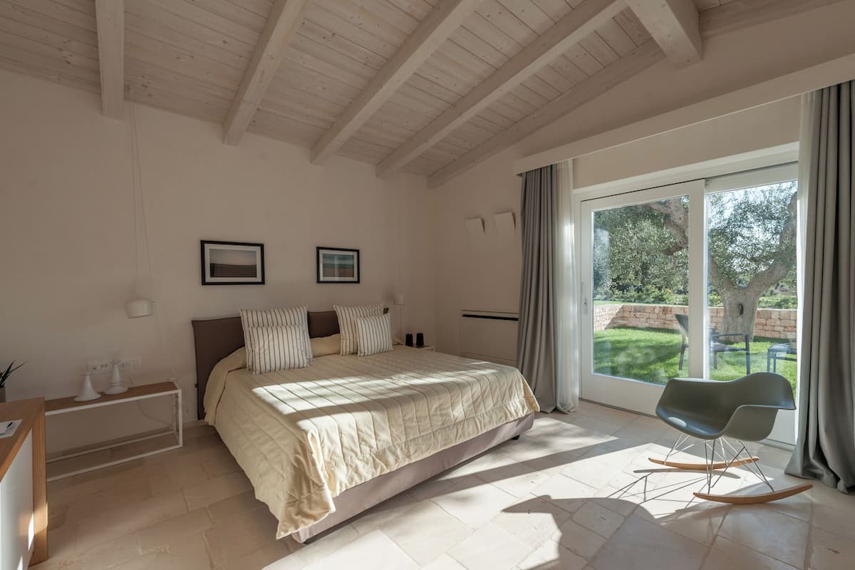 A bedroom with a large bed, a rocking chair, a desk, and a window with a view of the outdoors.