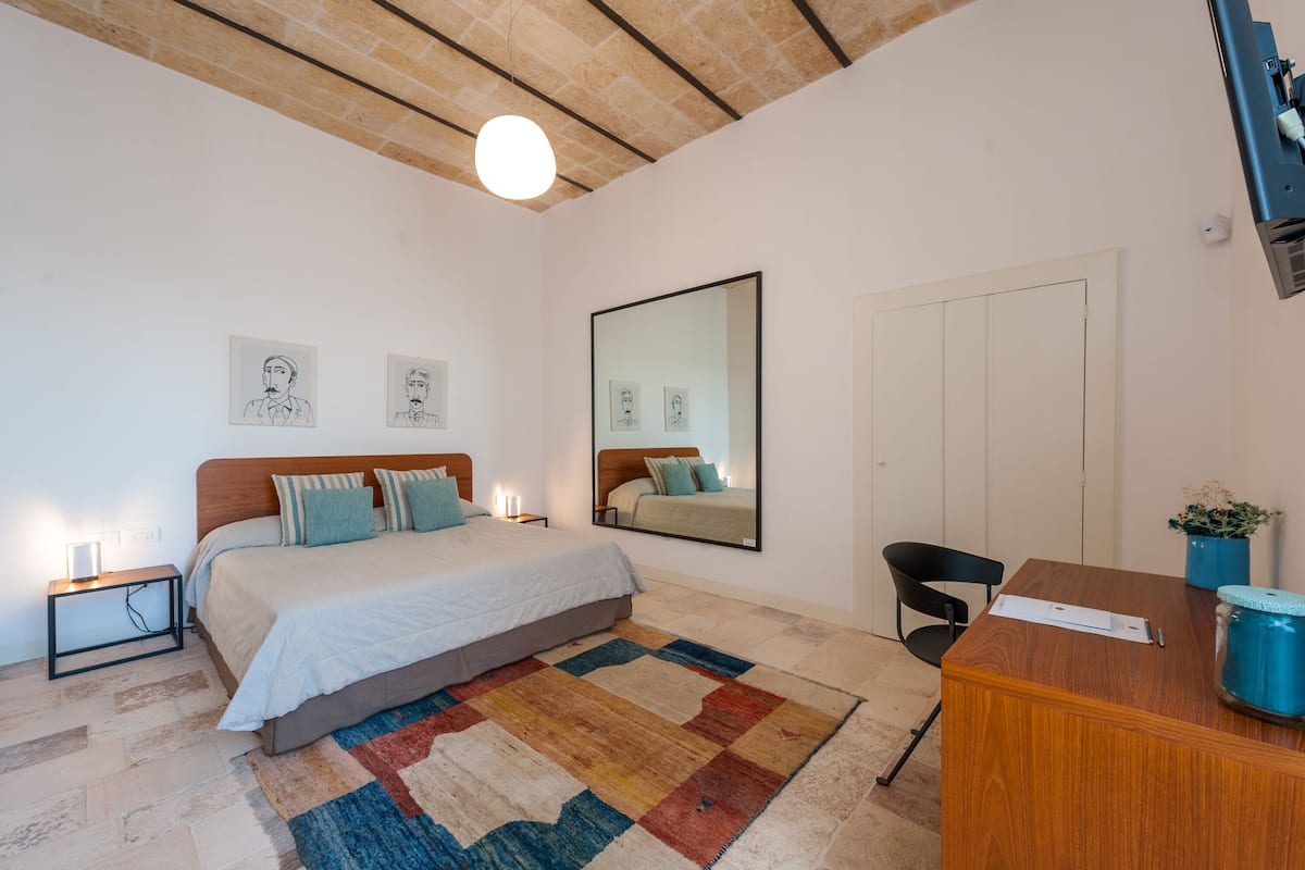 A bedroom with a bed, bedside tables, a desk, a chair, a mirror, and a door.