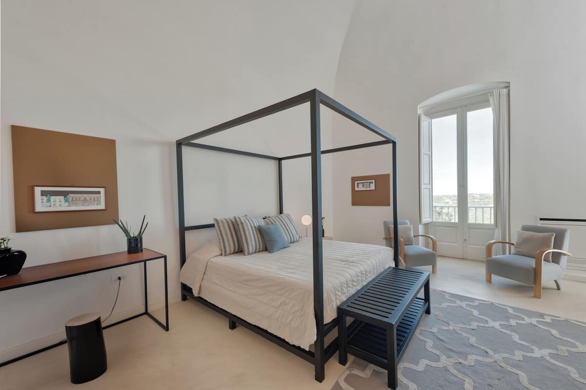 A modern bedroom with a four-poster bed, a desk, a chair, and a large window.