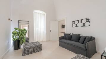 Un salón moderno con un sofá gris, una mesita, una planta en maceta y un gran ventanal con cortinas blancas.
