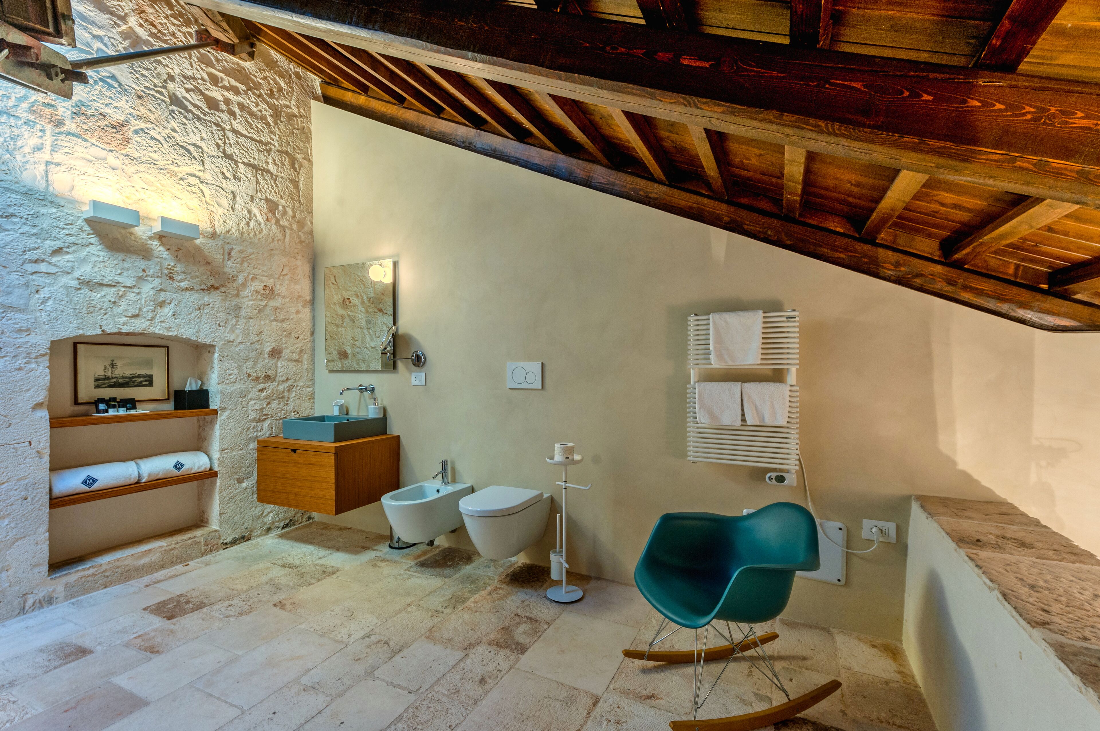Une salle de bain avec un mur en pierre, des poutres en bois, un lavabo, un bidet, un miroir et une chaise de couleur turquoise.