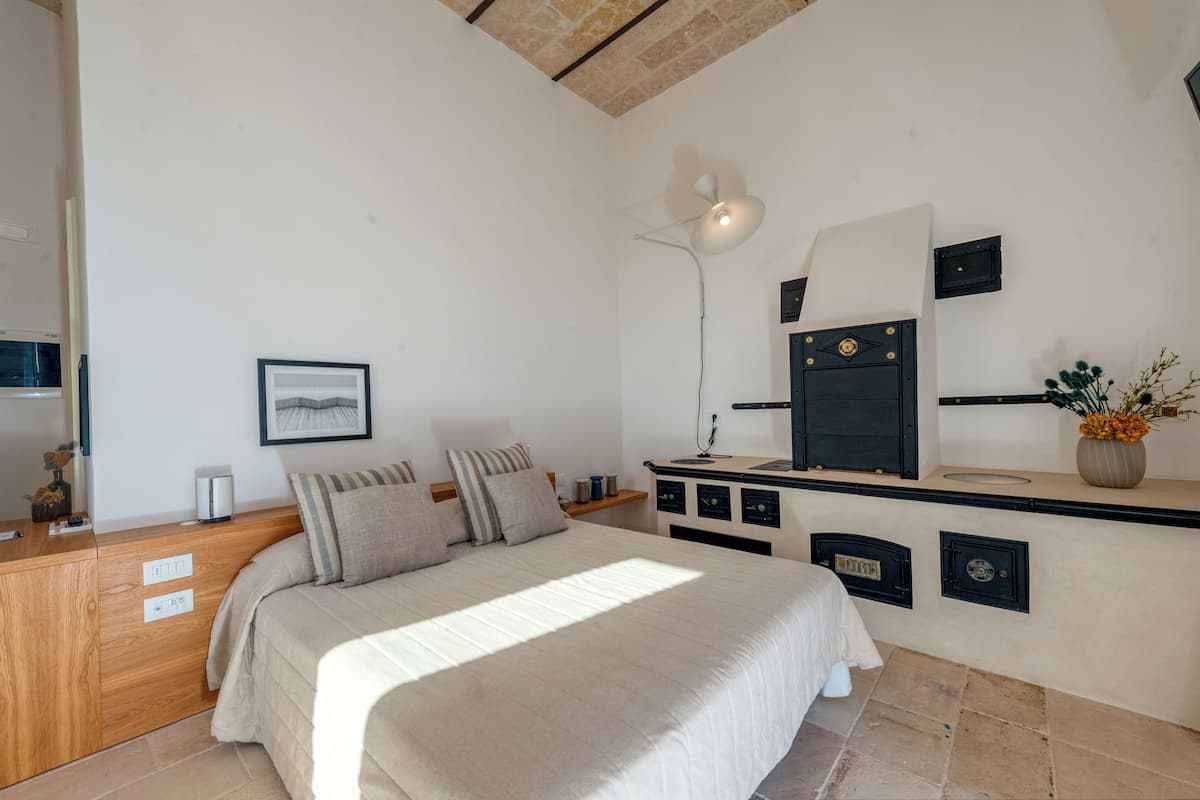 A bedroom with a bed, bedside table, lamp, and a small shelf with decorative items.