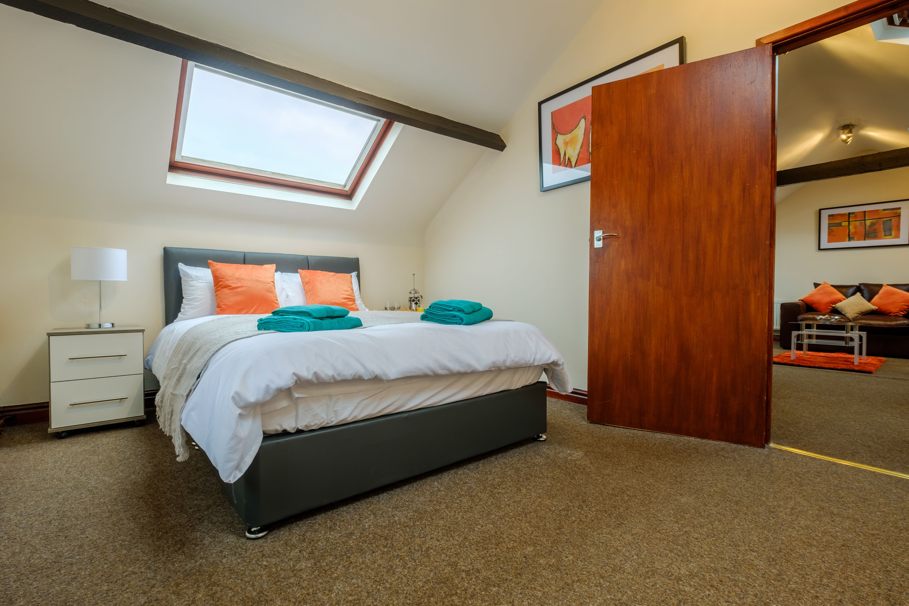 A bedroom with a skylight, a bed with teal and orange pillows, a bedside table with a lamp, and a wooden door leading to another room.
