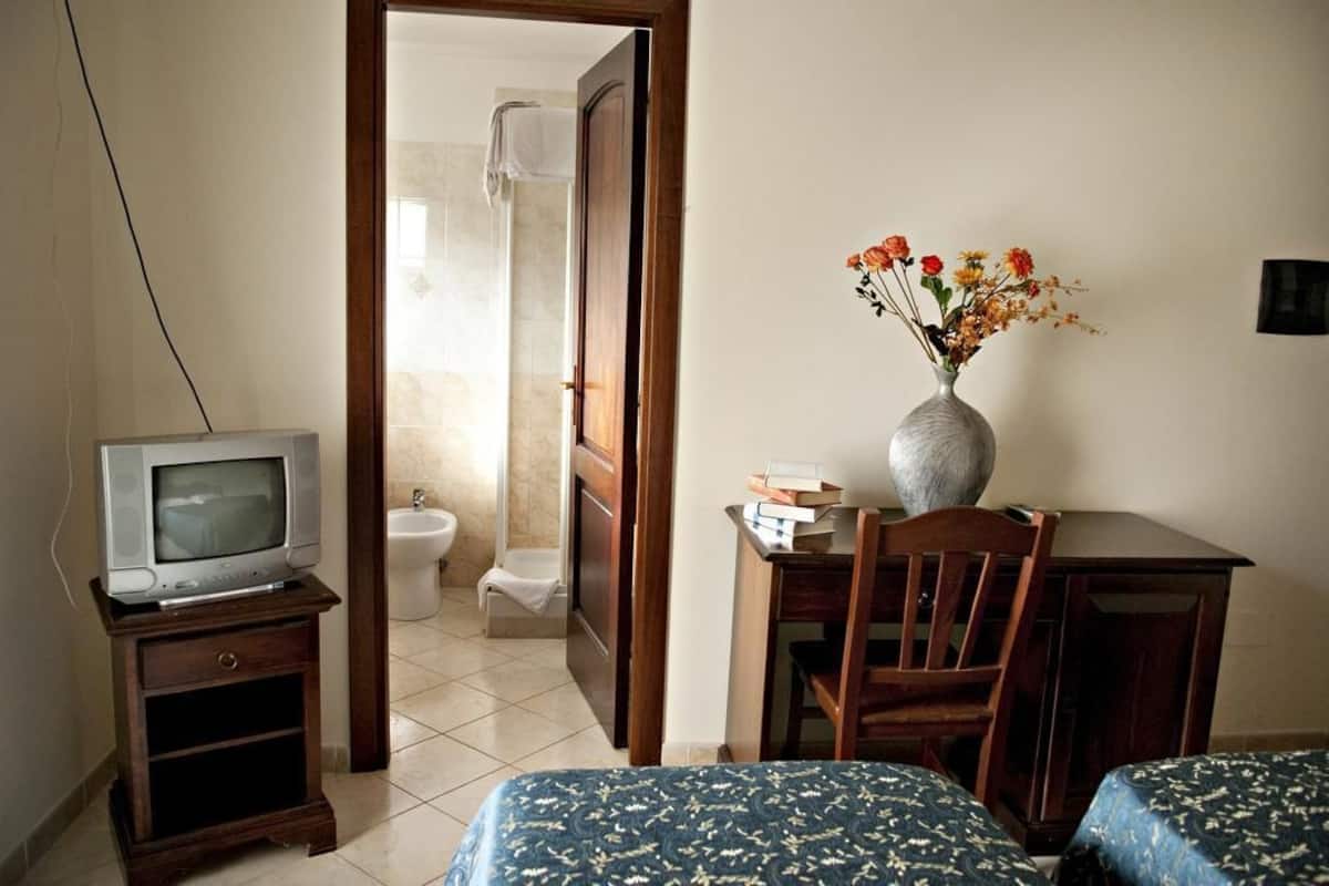 A hotel room with a TV on a wooden stand, a desk with a vase of flowers, a chair, and two beds with patterned bedding.