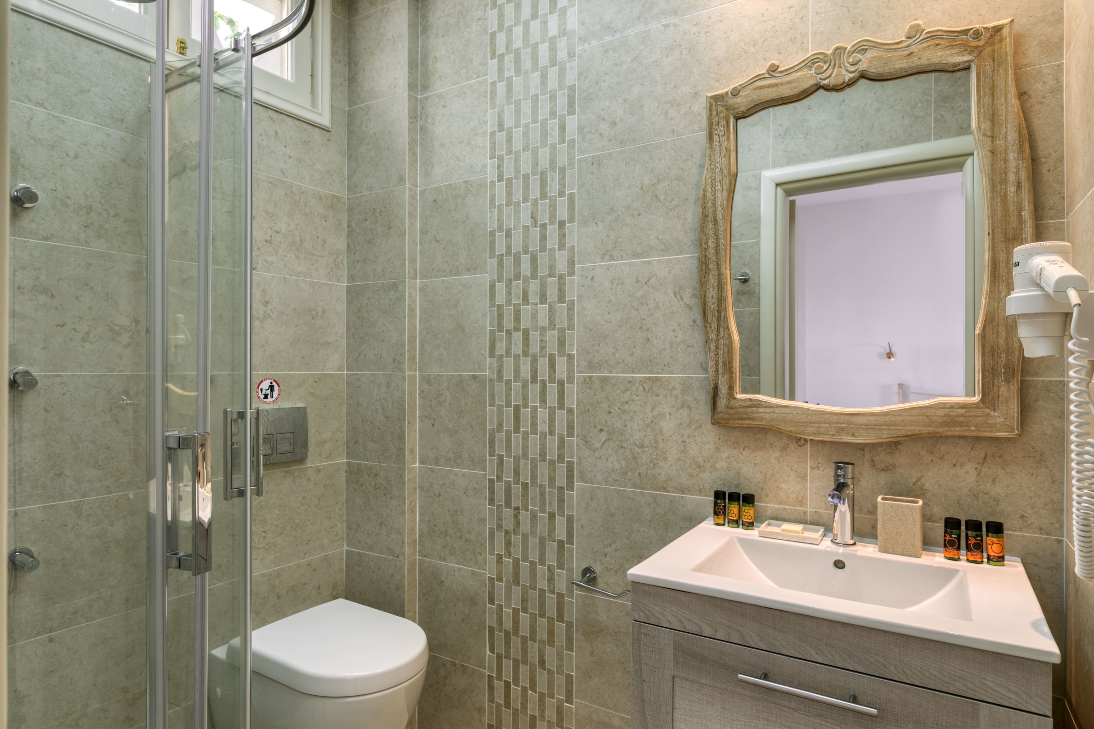 Un baño con cabina de ducha de vidrio, inodoro, lavabo con artículos de aseo personal y espejo con marco decorativo.