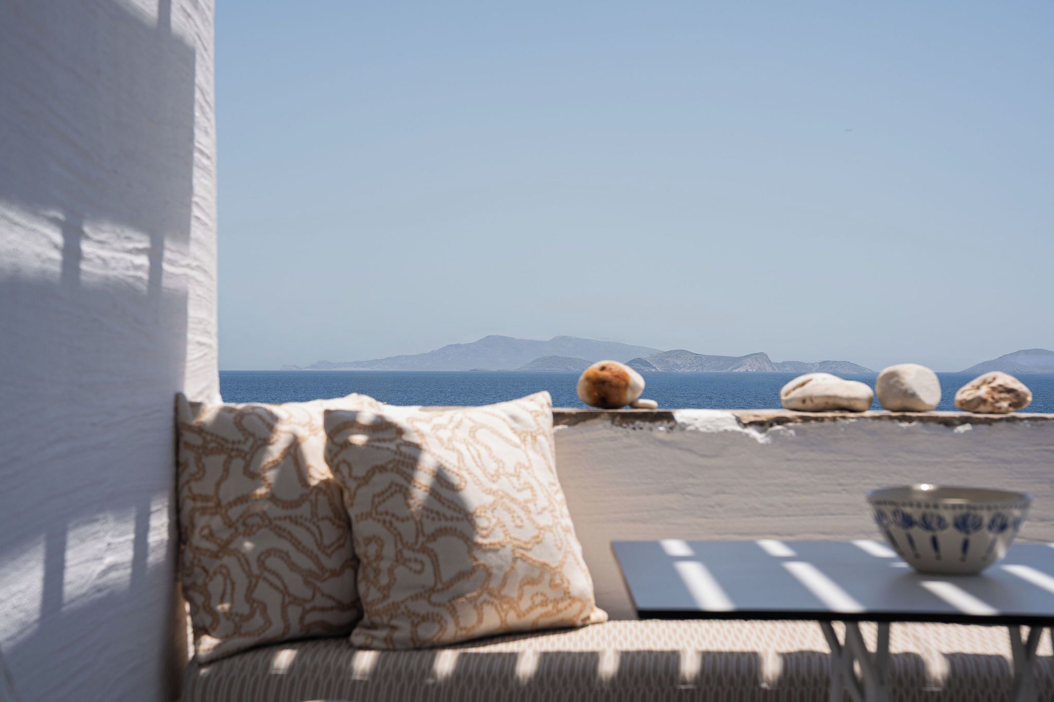 A balcony with a sofa, pillows, a bowl, and a view of the sea and mountains.