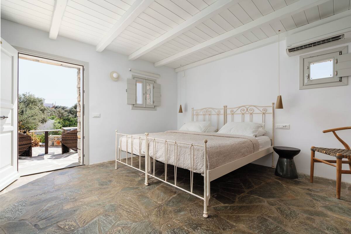 A bedroom with a stone floor, a bed with a metal frame, a wooden chair, and a small table.