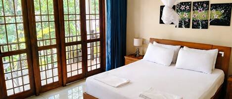 Habitación de hotel con cama, mesita de noche, lámpara, ventana con vista a follaje y una obra de arte en la pared.