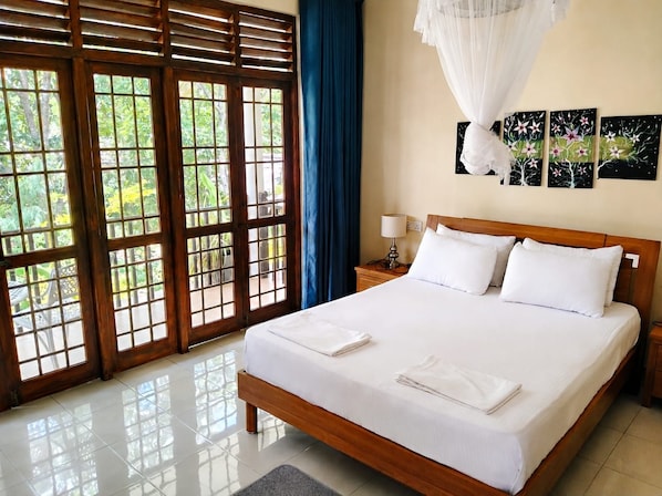 A hotel room with a bed, a nightstand, a lamp, a window with a view of greenery, and artwork on the wall.
