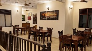 A dining area with multiple wooden tables and chairs, ceiling fans, wall-mounted artwork, and a staircase with a railing.