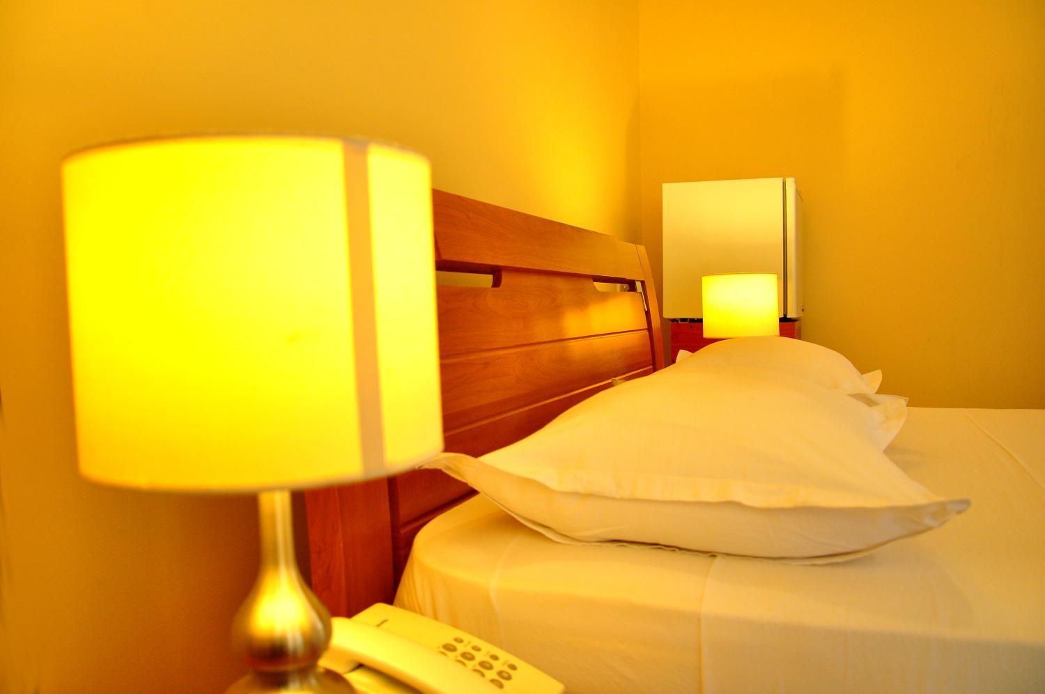 Een hotelkamer met een houten hoofdeinde, een bedlampje, een telefoon en een wit kussen op het bed.