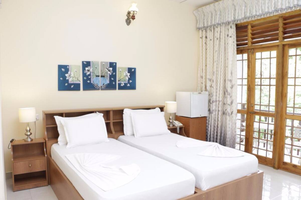 A hotel room with two beds, a wooden headboard, a nightstand with a lamp, a TV, and a large window with curtains.