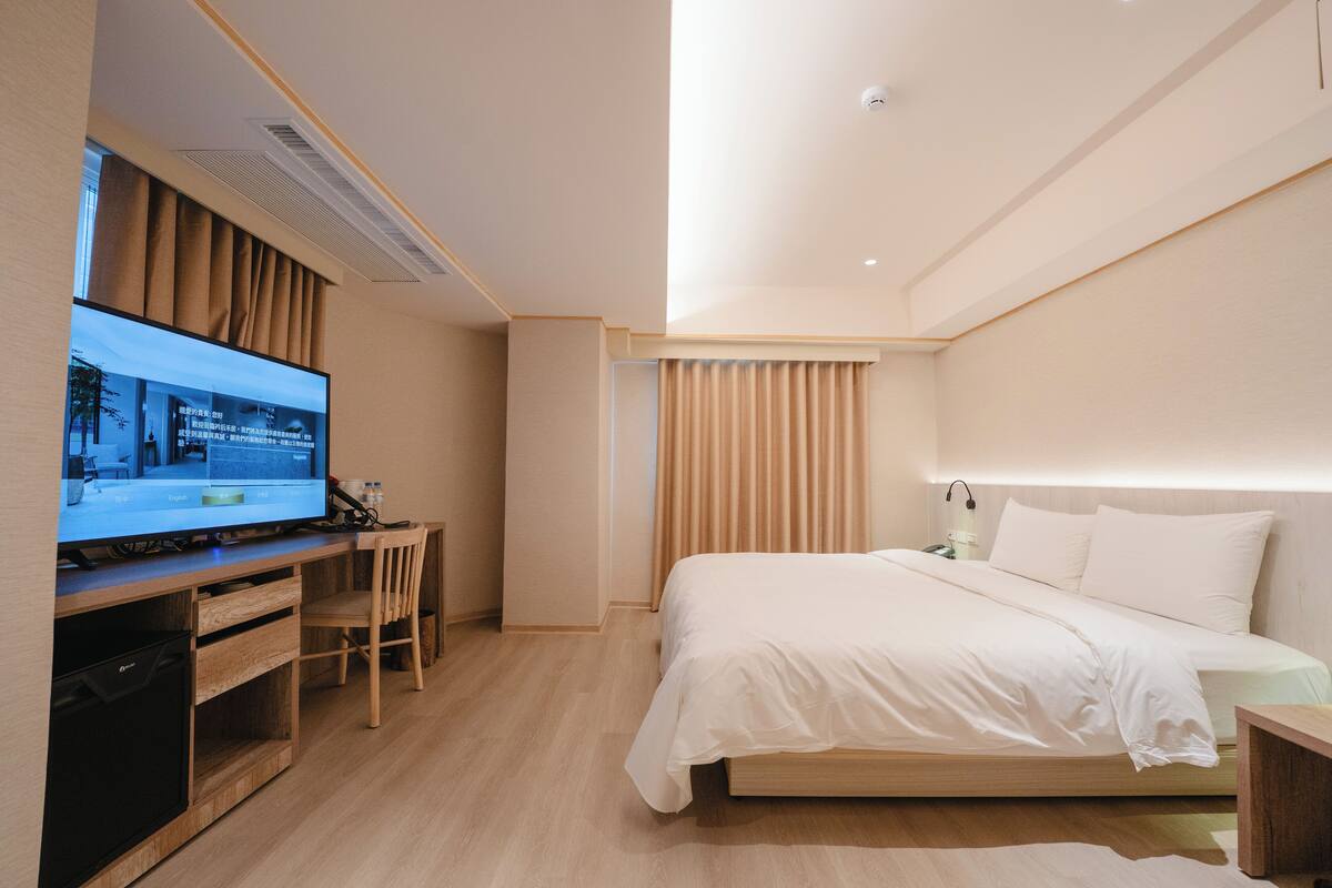 Ein modernes Hotelzimmer mit Bett, Schreibtisch mit Computer, Fernseher und einem Fenster mit Vorhängen.